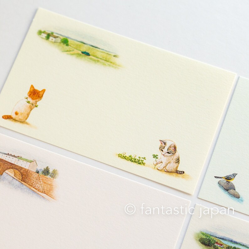 May include: A set of illustrated stationery featuring watercolor images. One card shows a ginger and white cat with a floral garland, another a gray and white cat with a four-leaf clover. Other cards depict landscapes and a bridge.