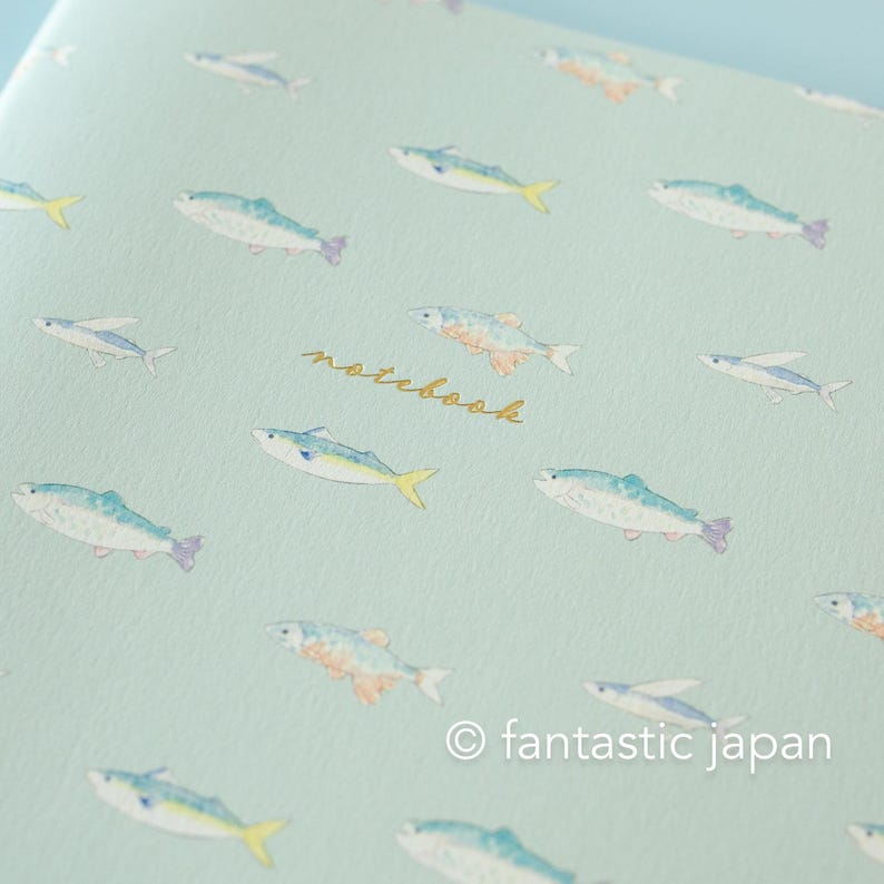 May include: A notebook with a light blue cover featuring a repeating pattern of watercolor-style fish in shades of blue, yellow, and orange. The word "notebook" is written in gold script. The copyright notice "fantastic japan" is at the bottom.