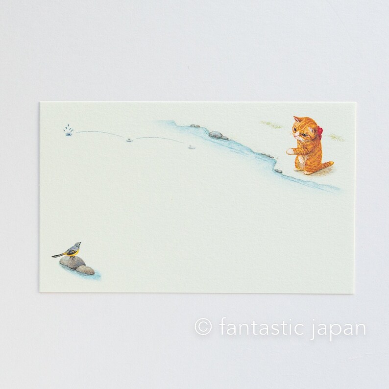 May include: Watercolor illustration of a ginger cat with a red bow, standing near a blue stream. A small bird sits on a rock in the foreground. The image has a light, airy feel with the text "fantastic japan" at the bottom.
