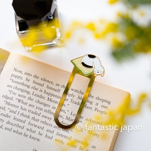 May include: Gold metal bookmark featuring a white cat with black spots sleeping on top of a closed book. The bookmark is on a page of an open book with text. The text reads "Now, into the silence, as the puppy, he found himself "Something else is happenin at Trade Mart. People are changing, Leader. Mento is."