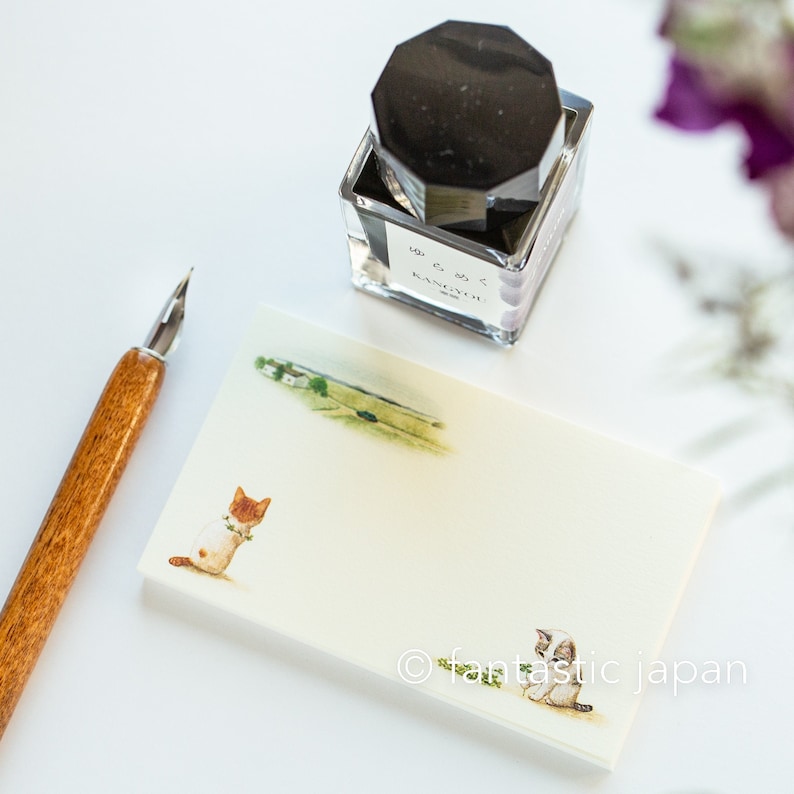 May include: A wooden-handled pen and a rectangular notepad with cat illustrations and a landscape scene. A clear glass ink bottle with a black cap sits above the notepad. The notepad features the text "fantastic japan".