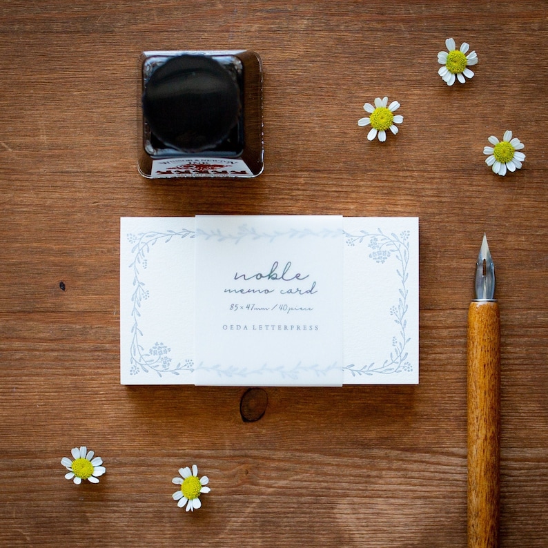 May include: A stack of white memo cards with a blue floral border and the text "noble memo card 85 x 47mm / 40 pieces OEDA LETTERPRESS" printed on the front. The cards are sitting on a wooden surface with a small bottle of ink and a calligraphy pen.