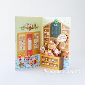 May include: A pop-up Christmas card depicting a festive bakery scene. The card features a red door, shelves filled with cakes, and Santa Claus figures. The words "Merry Christmas" are in a speech bubble.