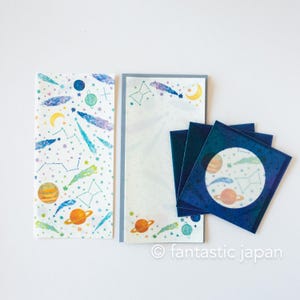 May include: Stationery set featuring a celestial theme. The set includes a notepad, a long sheet of paper, and several square cards. The designs feature planets, stars, constellations, and comets in shades of orange, blue, and yellow against a white background.