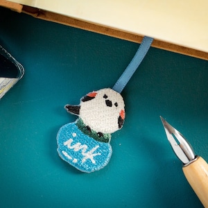 May include: Embroidered bookmark featuring a bird-like design with the word "ink" on a blue base. The bookmark is attached to a blue ribbon and is placed in a book. A bottle of ink and a calligraphy pen are also visible.