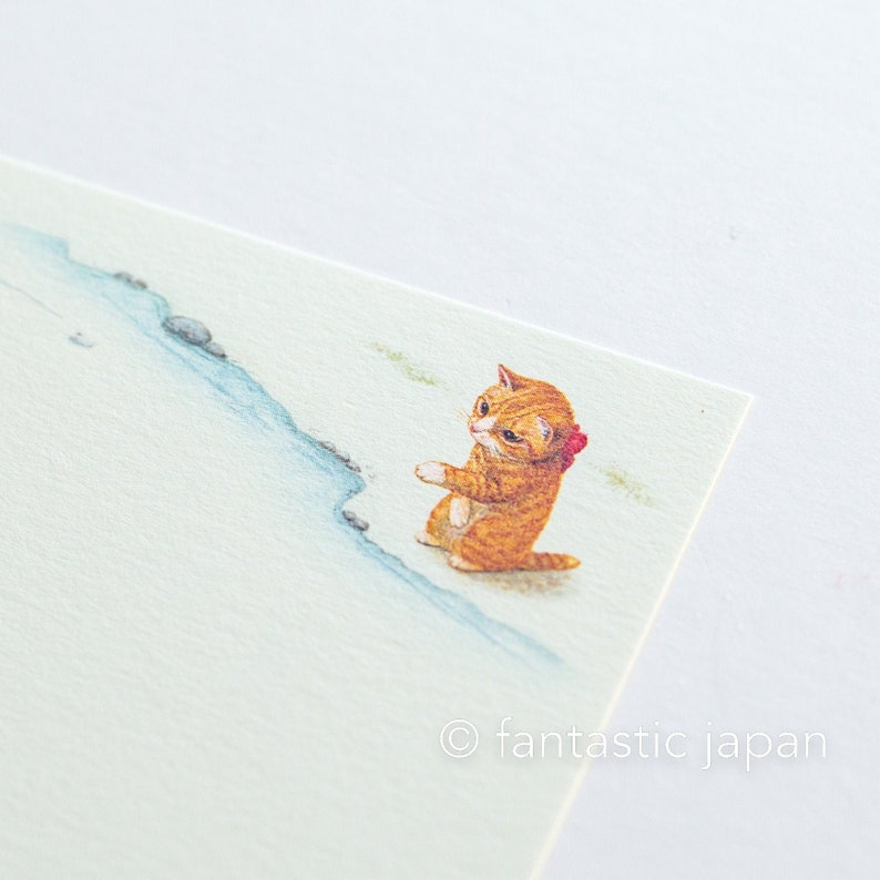 May include: A light green stationery card featuring an illustration of a ginger cat with a red bow. The cat is sitting near a blue stream with small rocks. The text "fantastic japan" is in the bottom right corner.