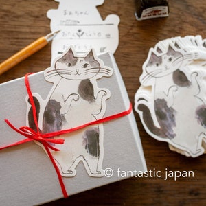 May include: A white gift box with a red ribbon tied around it. A black and white cat sticker is on the box. The sticker has a white background and a black cat with white markings. The cat is standing on its hind legs with its paws raised. The cat has a black nose and whiskers. The cat is looking to the right. The text "© fantastic japan" is printed on the box.