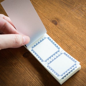 Puede incluir: Un peque&ntilde;o bloc de notas blanco con bordes decorativos azules. El bloc de notas est&aacute; abierto y la p&aacute;gina superior est&aacute; siendo volteada por una mano.