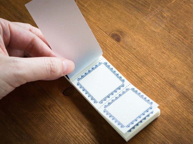 Puede incluir: Un peque&ntilde;o bloc de notas blanco con bordes decorativos azules. El bloc de notas est&aacute; abierto y la p&aacute;gina superior est&aacute; siendo volteada por una mano.