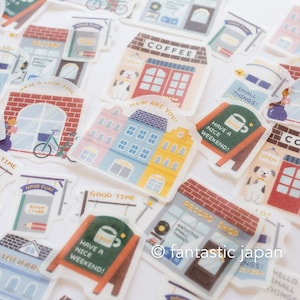 Op de afbeelding: Een verzameling kleurrijke stickers met illustraties van winkels en gebouwen. De stickers bevatten teksten als "Coffee", "Record Shop" en "Have a Nice Weekend!". De ontwerpen tonen verschillende architecturale stijlen en decoratieve details.