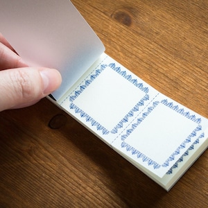 May include: A small notepad with blue decorative borders on each page. The notepad is open and the top page is being flipped back.