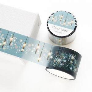 May include: Decorative clear tape with a sparkling design featuring white and gold starbursts on a teal background. The tape roll is next to a white box. The tape is labeled "clear tape - sparkler".