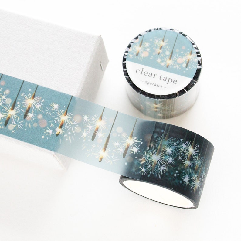 May include: Decorative clear tape with a sparkling design featuring white and gold starbursts on a teal background. The tape roll is next to a white box. The tape is labeled "clear tape - sparkler".