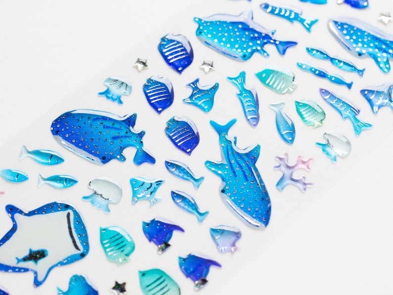 May include: A collection of blue and white glass fish and whale figurines on a white background. The figurines are all different shapes and sizes, and some have glitter accents.