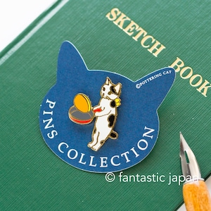 May include: A blue and white enamel pin featuring a black and white cat holding a red and yellow frying pan with a yellow egg inside. The pin is on a blue card with the text "PINS COLLECTION" and "POTTERING CAT" in white. The card is on a green surface.