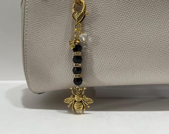 May include: A gold-toned bag charm featuring a heart-shaped lobster clasp, a small bee charm, a clear crystal bead, and three black faceted beads separated by two rhinestone-encrusted spacers, ending with a large gold-toned bee pendant.