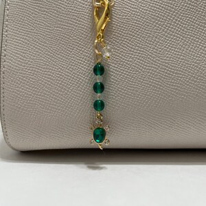 May include: A gold-colored bag charm with a lobster clasp, featuring green and clear beads, and a turtle pendant with a green gemstone and clear accents. The charm is attached to a light-colored handbag.