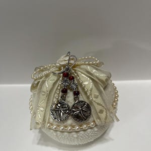 May include: A pair of silver-toned earrings featuring circular charms with embossed dragonfly designs, accented by small red beads and silver flower-shaped spacers, resting on a white textured sphere decorated with cream ribbon and pearl trim.