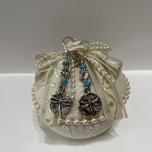 May include: A pair of silver-toned drop earrings featuring a circular charm with a dragonfly motif. Each earring includes a small, bright blue bead and a decorative silver spacer bead suspended from a simple silver-toned ear wire hook.