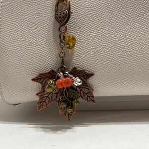 May include: A copper-toned metal bag charm featuring a large maple leaf pendant, a small orange pumpkin bead, and tiny acorn charms. The charm hangs from a heart-shaped lobster clasp and includes a single translucent yellow bead on a chain.