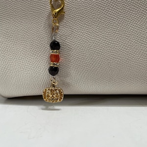 May include: A gold-toned lobster clasp bag charm featuring a vertical arrangement of two black faceted beads, one orange bead, and two small rhinestone-encrusted spacers, ending with a gold-toned pumpkin charm covered in small clear crystals.