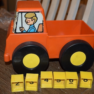 Könnte beinhalten: Ein orangefarbener Spielzeug-LKW aus Kunststoff mit schwarzen Rädern und gelben Zentren. Der LKW hat eine Fahrerillustration im Fenster. Enthalten sind gelbe Blöcke mit den Zahlen 2, 3, 4, 5, 6 und 0. Ein klassisches Kinderspielzeug.