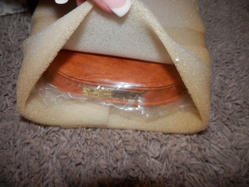 May include: A wooden oval-shaped box with a gold plaque that reads "Celebrating Five Cherished Years". The box is wrapped in clear plastic and surrounded by white foam padding.