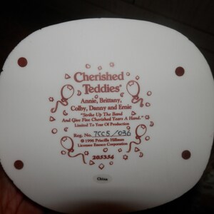 May include: White ceramic figurine base with a red inscription that reads "Cherished Teddies" with the names Annie, Brittany, Colby, Danny and Ernie. The inscription also reads "Strike Up The Band And Give Five Cherished Years A Hand." The base also includes the registration number 7CC5/036, the year 1996, the name Priscilla Hillman, the company Enesco Corporation, and the number 205354. The base is made in China.