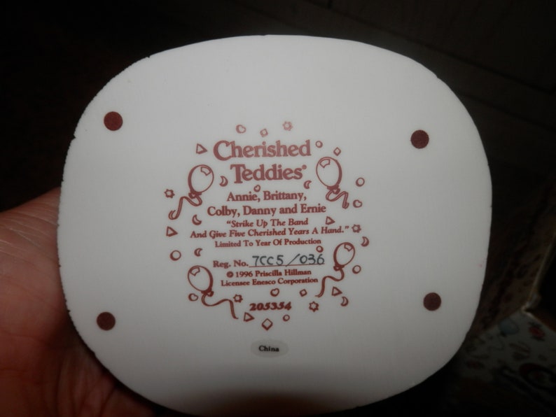 May include: White ceramic figurine base with a red inscription that reads "Cherished Teddies" with the names Annie, Brittany, Colby, Danny and Ernie. The inscription also reads "Strike Up The Band And Give Five Cherished Years A Hand." The base also includes the registration number 7CC5/036, the year 1996, the name Priscilla Hillman, the company Enesco Corporation, and the number 205354. The base is made in China.