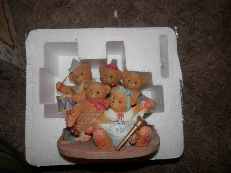 May include: A group of five brown teddy bears wearing party hats and playing musical instruments. The bears are arranged on a wooden base with a banner that reads "Celebrate".
