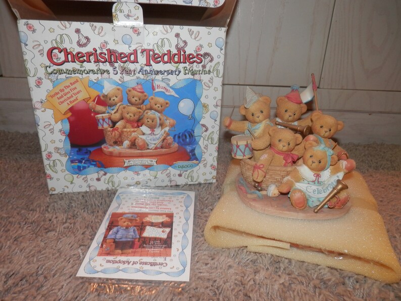 May include: A group of five teddy bears dressed in party hats and outfits are playing musical instruments in a basket. The bears are part of a Cherished Teddies commemorative 5 year anniversary figurine. The figurine is on a brown and white base with the words "Celebrate" and "Cherished Years" on it.
