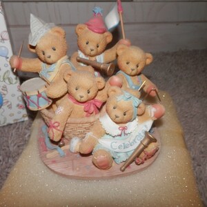 May include: Five brown teddy bears wearing party hats and playing musical instruments. The bears are sitting in a basket and are surrounded by confetti and streamers. The text "Celebrate" is written on the front of the basket.
