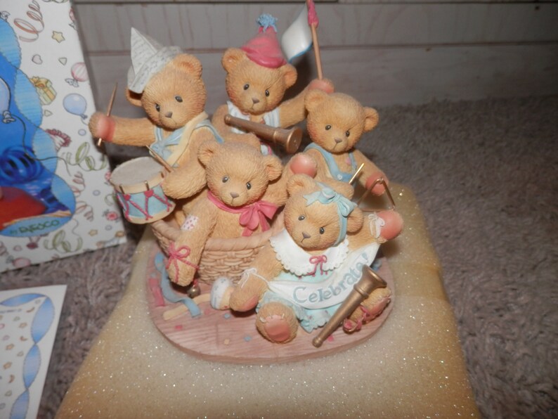 May include: Five brown teddy bears wearing party hats and playing musical instruments. The bears are sitting in a basket and are surrounded by confetti and streamers. The text "Celebrate" is written on the front of the basket.