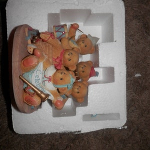 May include: A group of five brown teddy bears playing musical instruments. The bears are wearing colorful outfits and are sitting on a wooden platform. The platform is surrounded by white foam packaging. The bears are playing a trumpet, a drum, a tambourine, a cymbal, and a horn. The text "Celebrate" is visible on the platform.