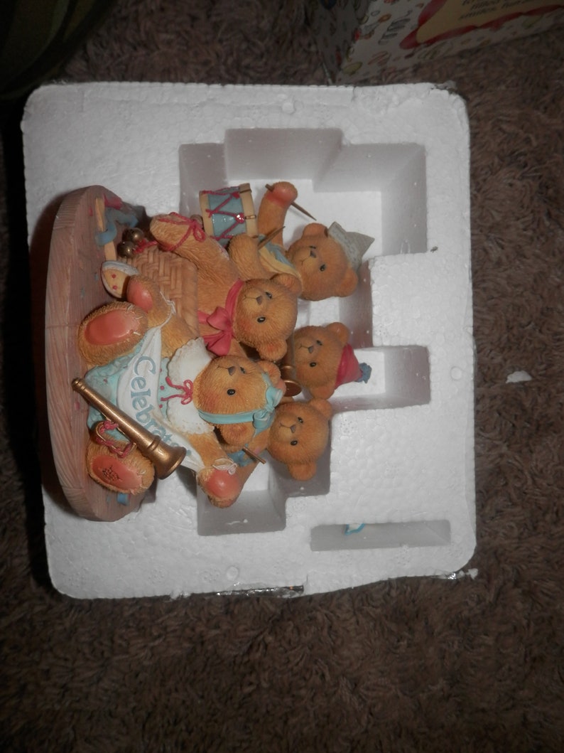 May include: A group of five brown teddy bears playing musical instruments. The bears are wearing colorful outfits and are sitting on a wooden platform. The platform is surrounded by white foam packaging. The bears are playing a trumpet, a drum, a tambourine, a cymbal, and a horn. The text "Celebrate" is visible on the platform.