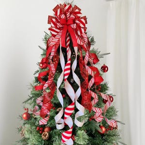 May include: A Christmas tree topper featuring a large, multi-layered bow made of red and white striped ribbon, solid red ribbon, and sheer red ribbon with gold patterns. Long, cascading ribbon tails drape down the green pine tree branches.