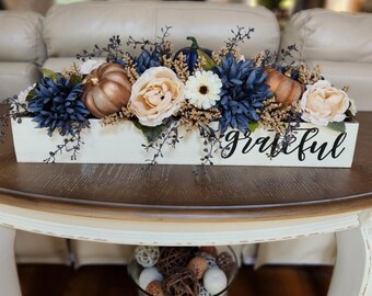 May include: A decorative centerpiece featuring a white wooden box with the word "Grateful" in black script. The box is filled with blue and cream-colored flowers, bronze pumpkins, and greenery. The arrangement sits on a brown and white table.
