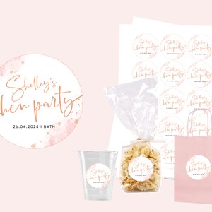 May include: A pink and gold round sticker with the text "Shelley's hen party 26.04.2024 | BATH" in a script font. The sticker has a watercolor background with pink and gold splatters.