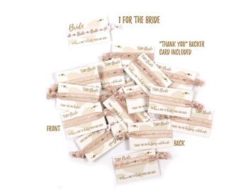 May include: A stack of pink and gold hair ties with a white card attached. The card has the text "Team Bride" and "To have and to hold your hair back". The back of the card says "Thank you for helping celebrate".