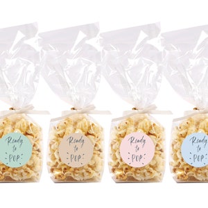 Puede incluir: Cuatro bolsas de celofán transparentes llenas de palomitas de maíz, cada una atada con una cinta dorada y una pegatina redonda que dice "Ready to POP" en diferentes colores: azul, verde, rosa y beige.