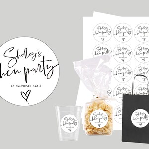 May include: A set of black and white round stickers with the text "Shelly's hen party 26.04.2024 | BATH" and a small heart. The stickers are on a white sheet of paper, a clear plastic bag, a clear plastic cup, a black paper bag, and a white paper bag.
