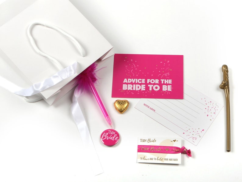 May include: A white gift bag with a pink feather pen, a pink and white button that says "Team Bride", a pink and white ribbon, a gold heart-shaped chocolate, and a pink card that says "Advice for the Bride to Be" with a white card attached that says "With Love."