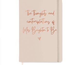 May include: A light pink notebook with a vertical elastic closure and a ribbon bookmark. The cover features copper-colored script text that reads The Thoughts and contemplations of Mrs Brighton To Be, accompanied by a small hand-drawn heart icon.