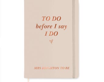 May include: A light pink notebook with a vertical elastic closure band. The cover features metallic copper text that reads TO DO before I say I DO, a small heart icon, and the personalized text MRS BRIGHTON TO BE at the bottom.