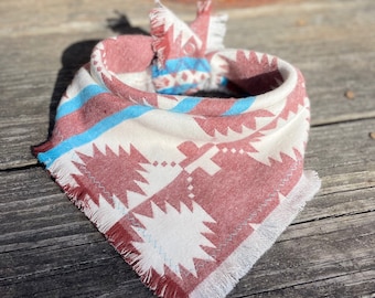 May include: A triangular pet bandana featuring a woven pattern in shades of dusty rose, cream, and bright turquoise. The fabric has a textured appearance with frayed edges and a knotted top, showcasing geometric shapes and cross motifs throughout.