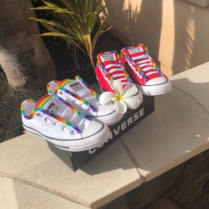May include: Two pairs of white Converse sneakers with rainbow and red beaded laces. The sneakers are on a black Converse box with the word "Converse" printed on it.