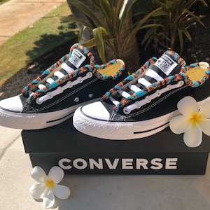 May include: A pair of black Converse sneakers with white soles and laces. The laces are decorated with colorful beaded trim. The shoes are sitting on a black box with the word "CONVERSE" printed on the front.