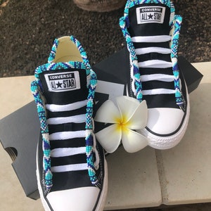 May include: A pair of black Converse All Star sneakers with blue, white, and turquoise beaded laces. The laces are tied in a criss-cross pattern and the shoes are sitting on a gray surface.