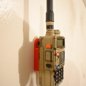 May include: A tan and black two-way radio with an orange button is mounted on a red wall mount. The radio has a black antenna and a keypad with multiple buttons.
