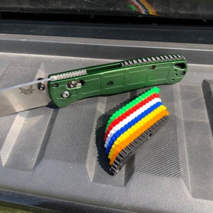 May include: A green pocket knife with a black handle and a butterfly logo on the blade. The knife is lying on a black textured surface with a stack of colorful plastic gear-shaped objects.